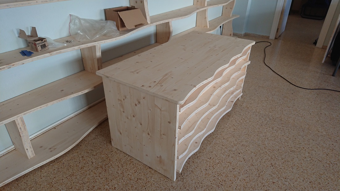 Mesa de madera con un frente de perfiles ondulados, situada junto a las estanterías de la biblioteca.